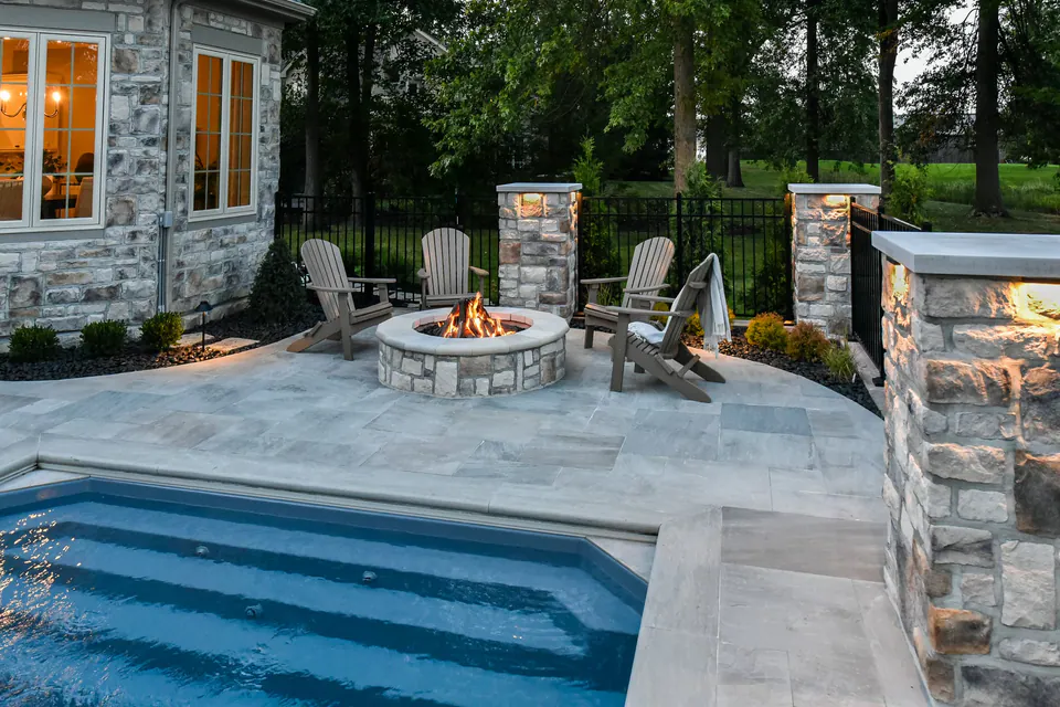 Pool steps beside paver deck, firepit seating, stone piers, and evening lighting